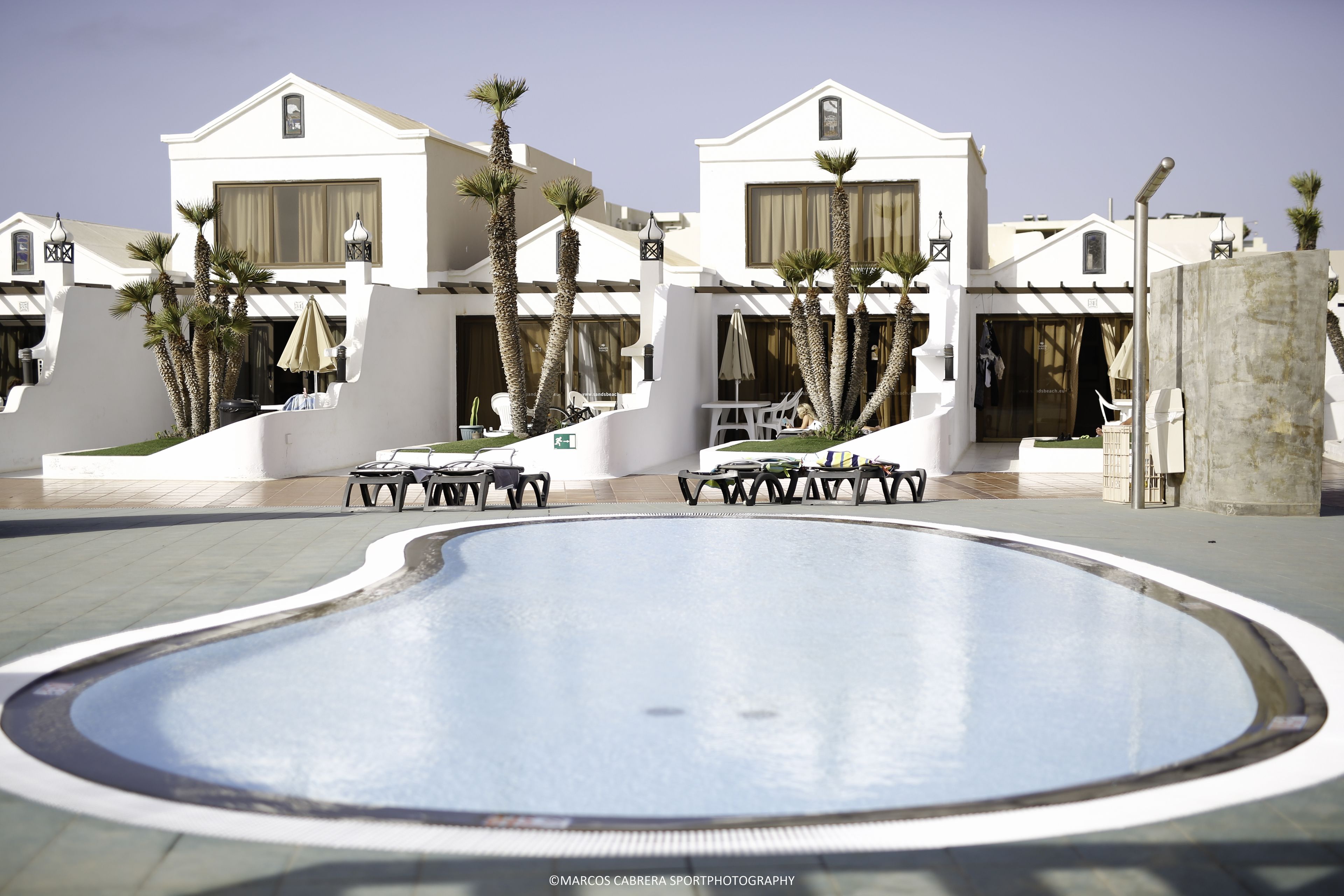 Una piscina con sillas de descanso y sombrillas, rodeada de edificios blancos con balcones y palmeras.