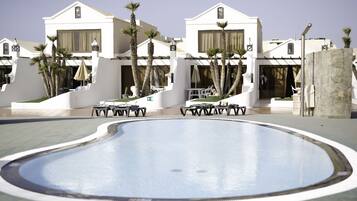 Una piscina con sillas de descanso y sombrillas, rodeada de edificios blancos con balcones y palmeras.
