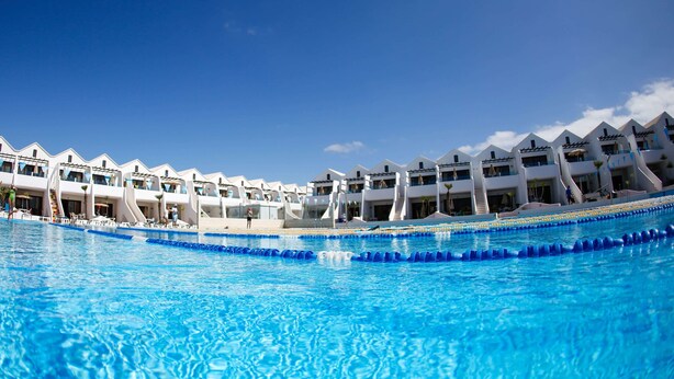 A swimming pool with clear blue water, surrounded by white buildings with balconies and a clear sky. - Sands Beach Resort (Teguise)