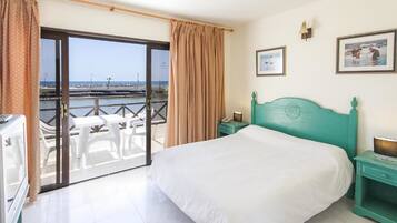 Habitación de hotel con cama, mesita de noche, televisor y balcón con sillas blancas y vista al mar.
