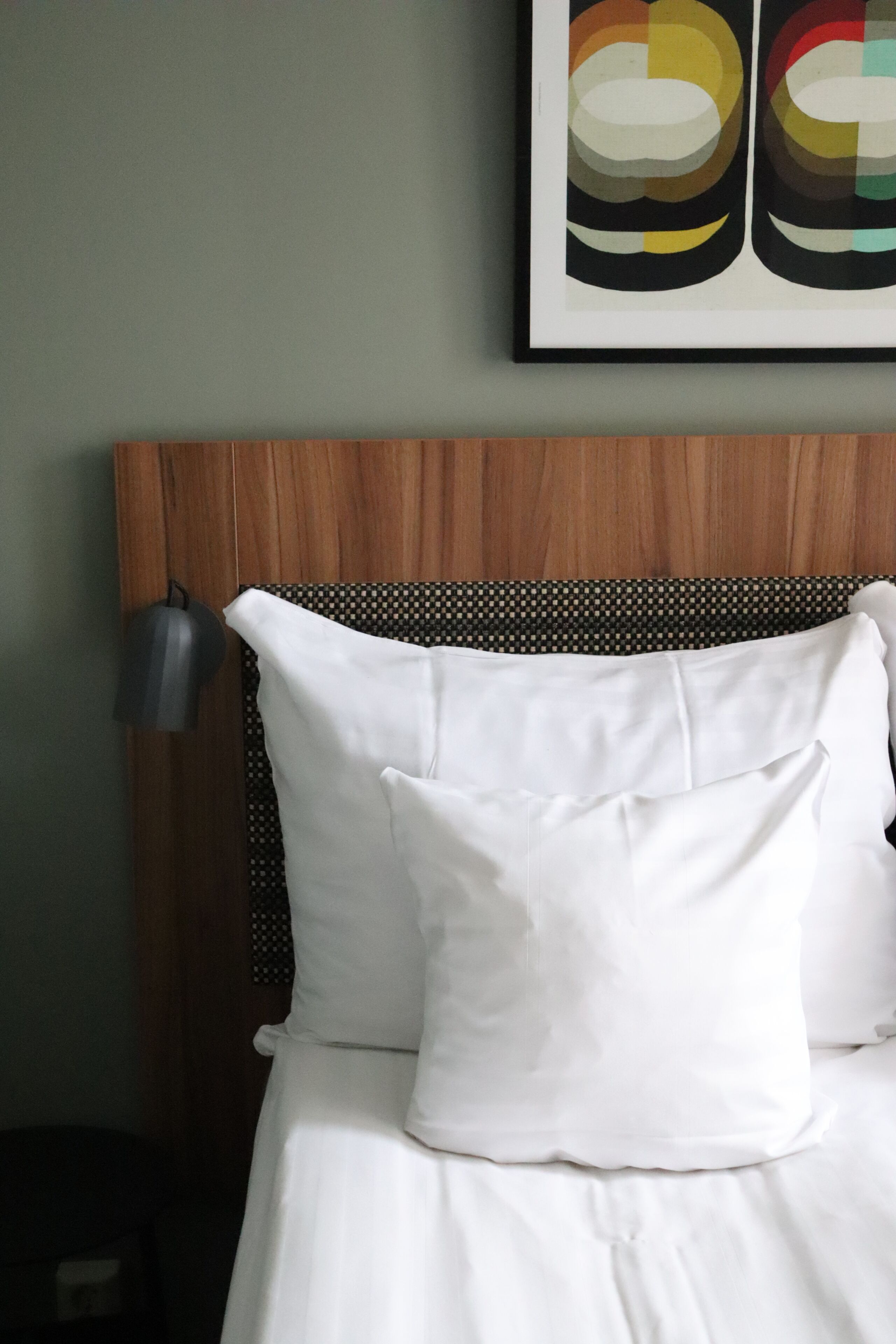 A bed with white bedding, a wooden headboard, and a wall-mounted lamp.