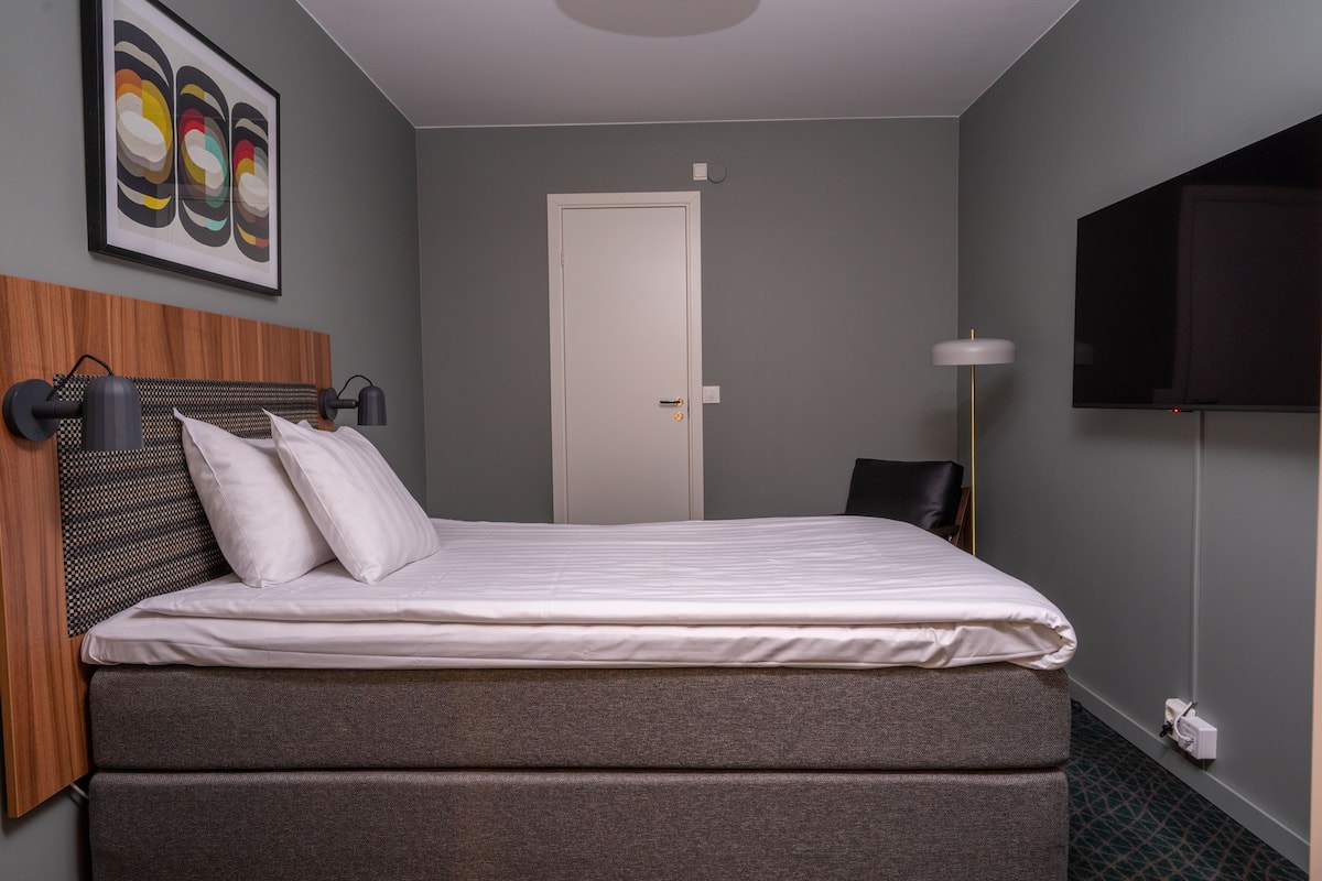 Una cama bien hecha con colchón gris y ropa de cama blanca, cabecera de madera, televisor montado en la pared y una obra de arte abstracto enmarcada sobre la cama.