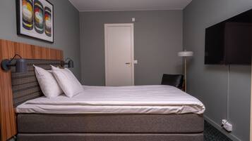 A neatly made bed with a grey mattress and white linens, a wooden headboard, a wall-mounted TV, and a framed abstract art piece above the bed.