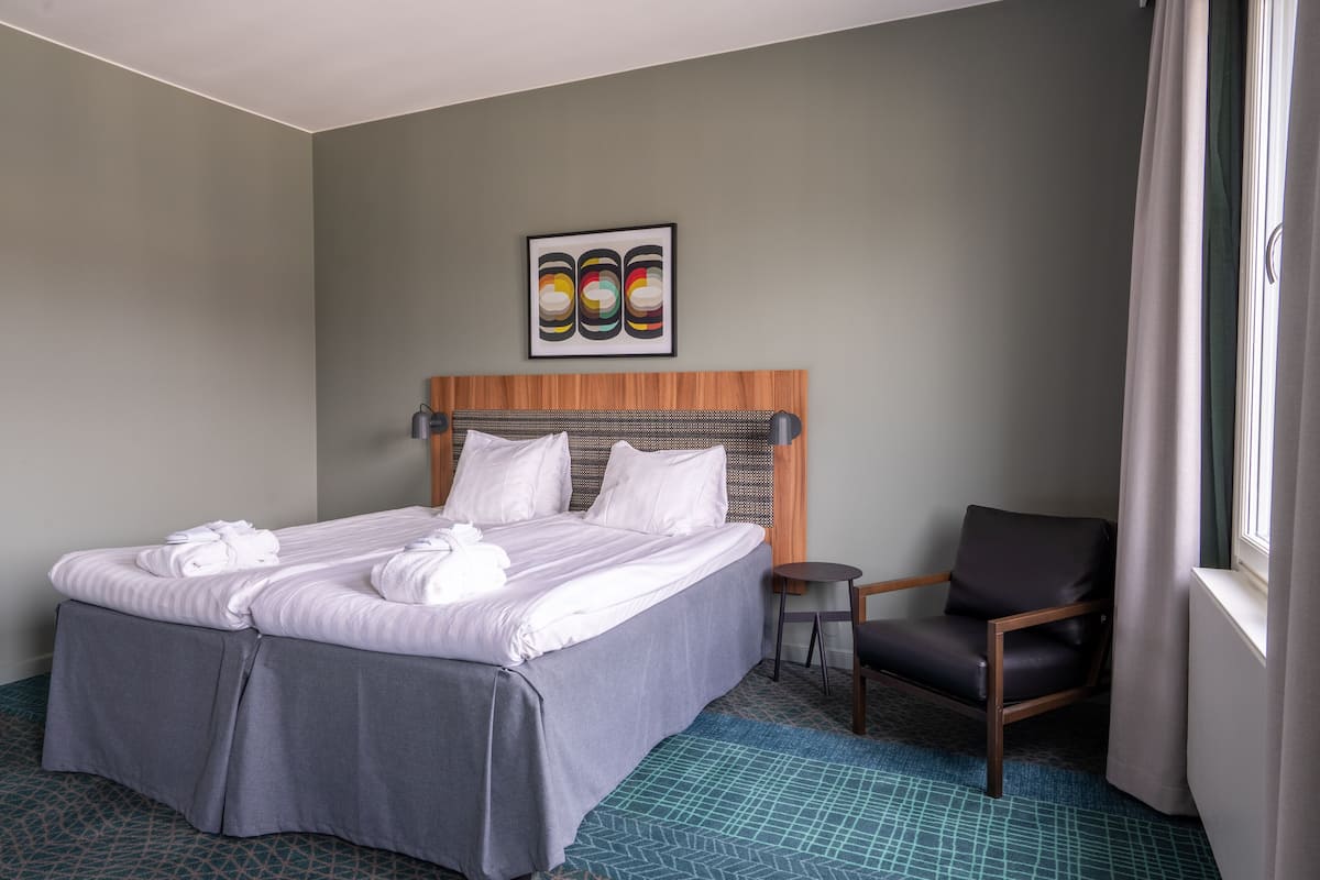 Habitación de hotel con una cama grande, cabecera de madera, una mesita, una silla y una ventana con cortinas.