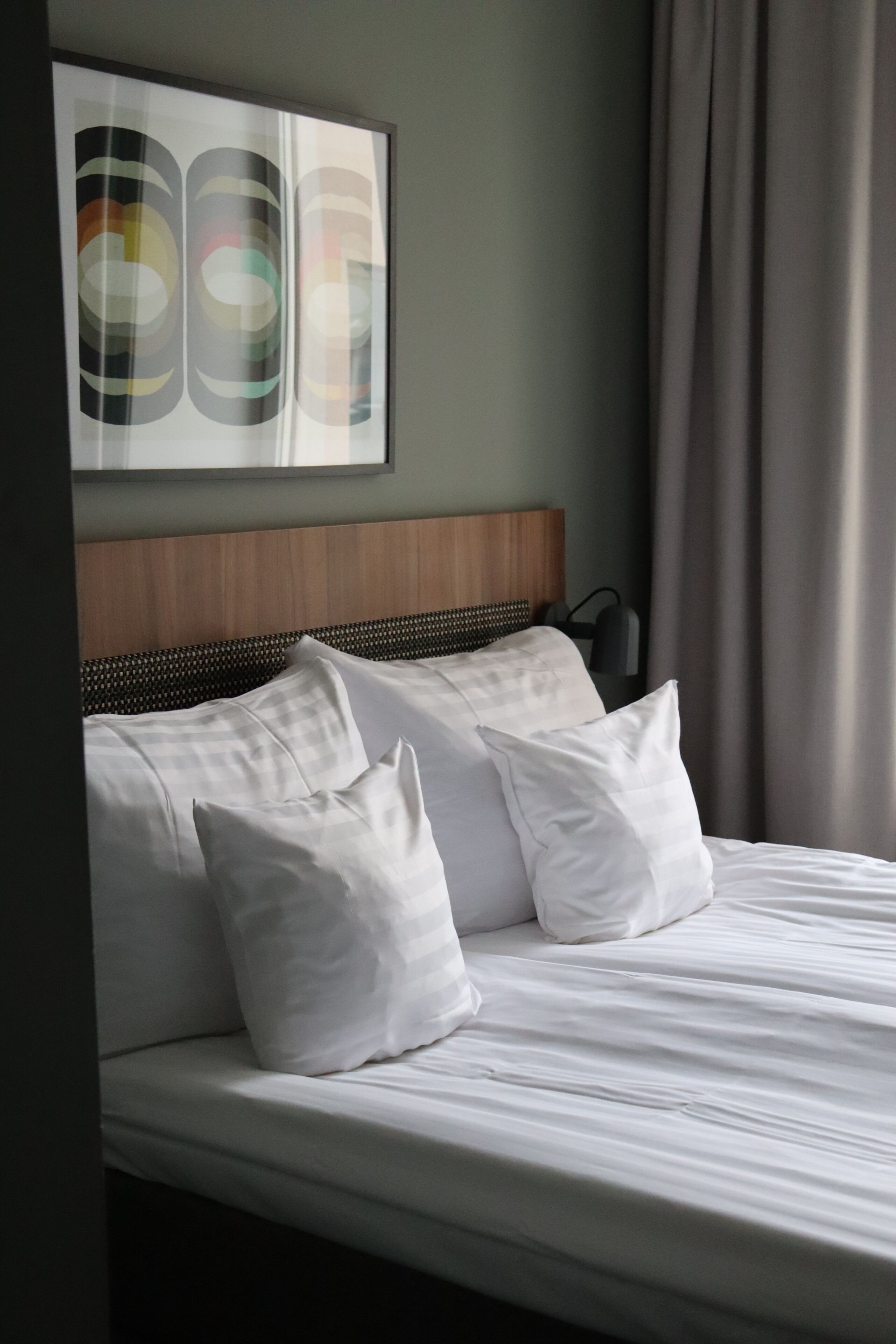 A neatly made bed with white bedding and pillows, a wooden headboard, and a wall-mounted abstract artwork.