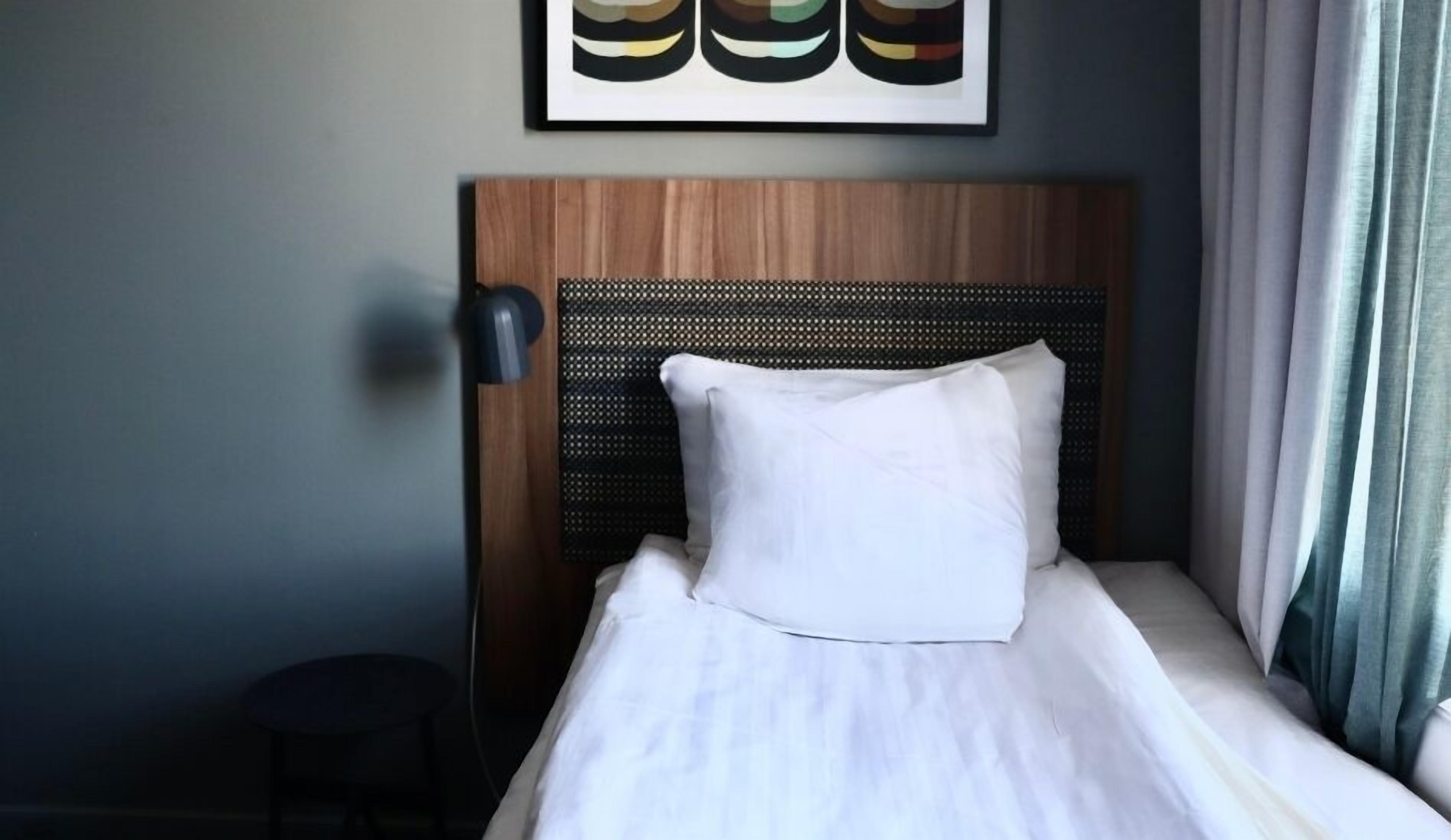 Una habitación de hotel con cabecera de madera, almohada blanca, mesita de noche pequeña y lámpara fijada a la pared.