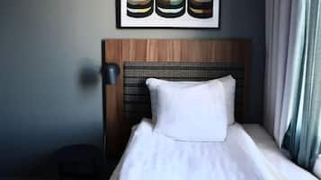 Una habitación de hotel con cabecera de madera, almohada blanca, mesita de noche pequeña y lámpara fijada a la pared.