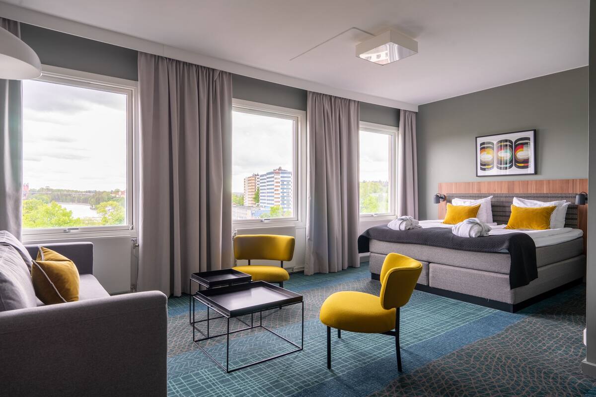 Una habitación de hotel con una cama, dos sillas amarillas, una mesita pequeña y ventanas grandes con cortinas.