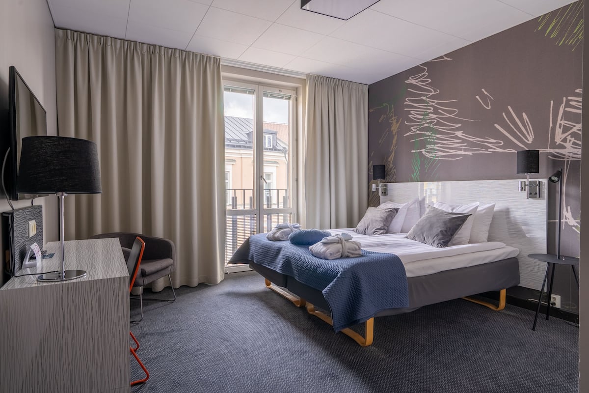Habitación de hotel con cama, escritorio, silla, televisor y ventana con cortinas.