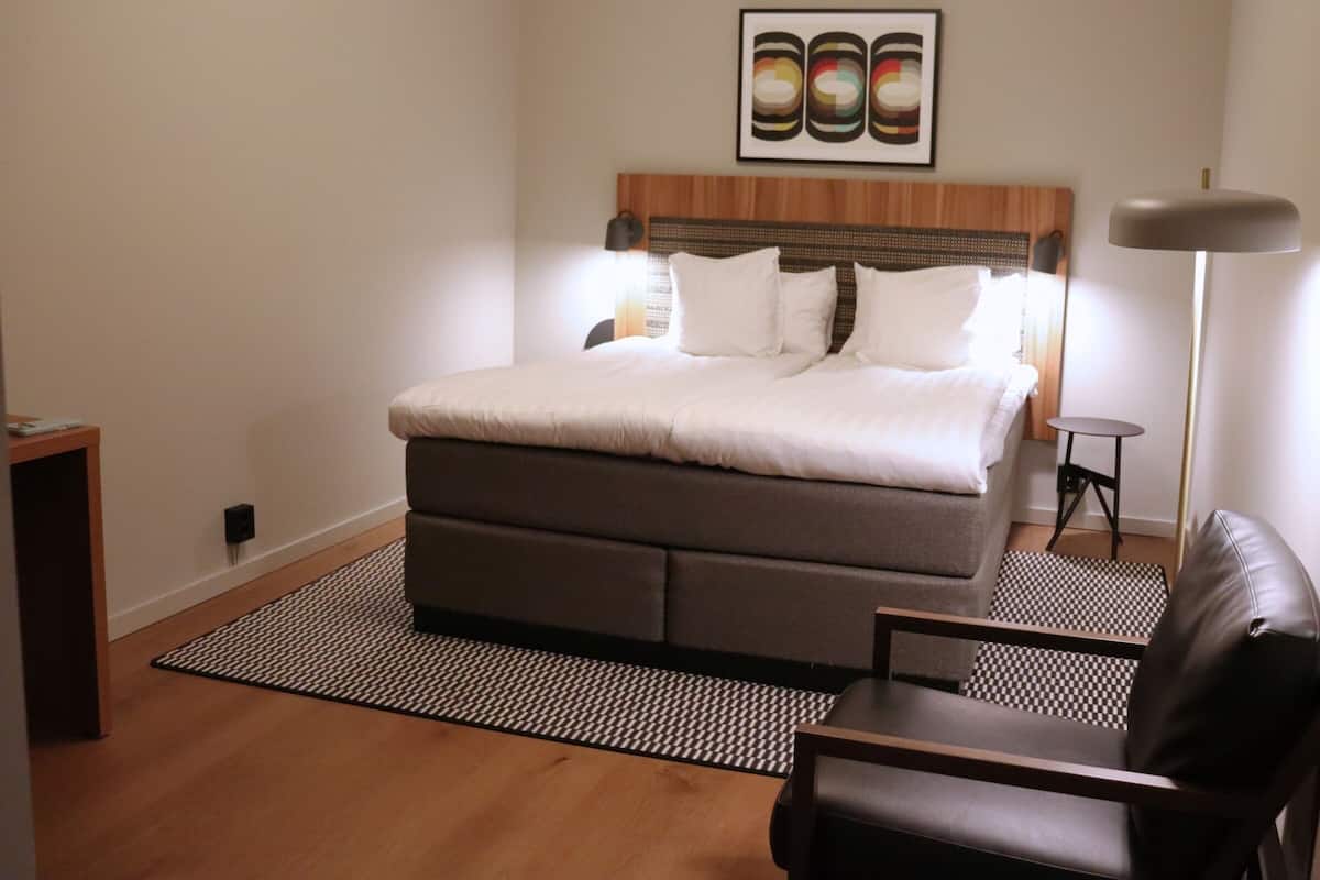 Un dormitorio con cama, mesita de noche, una silla, lámpara de pie y una obra de arte colgada en la pared.