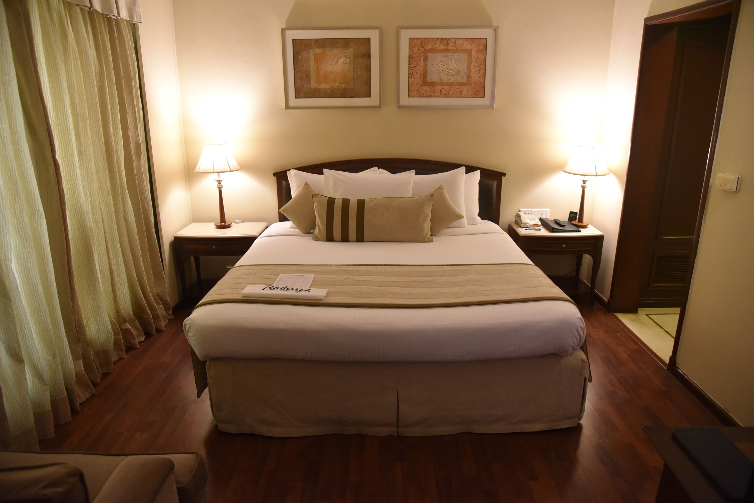 A hotel room with a large bed, two bedside tables with lamps, a nightstand, a wooden floor, and two framed pictures on the wall.