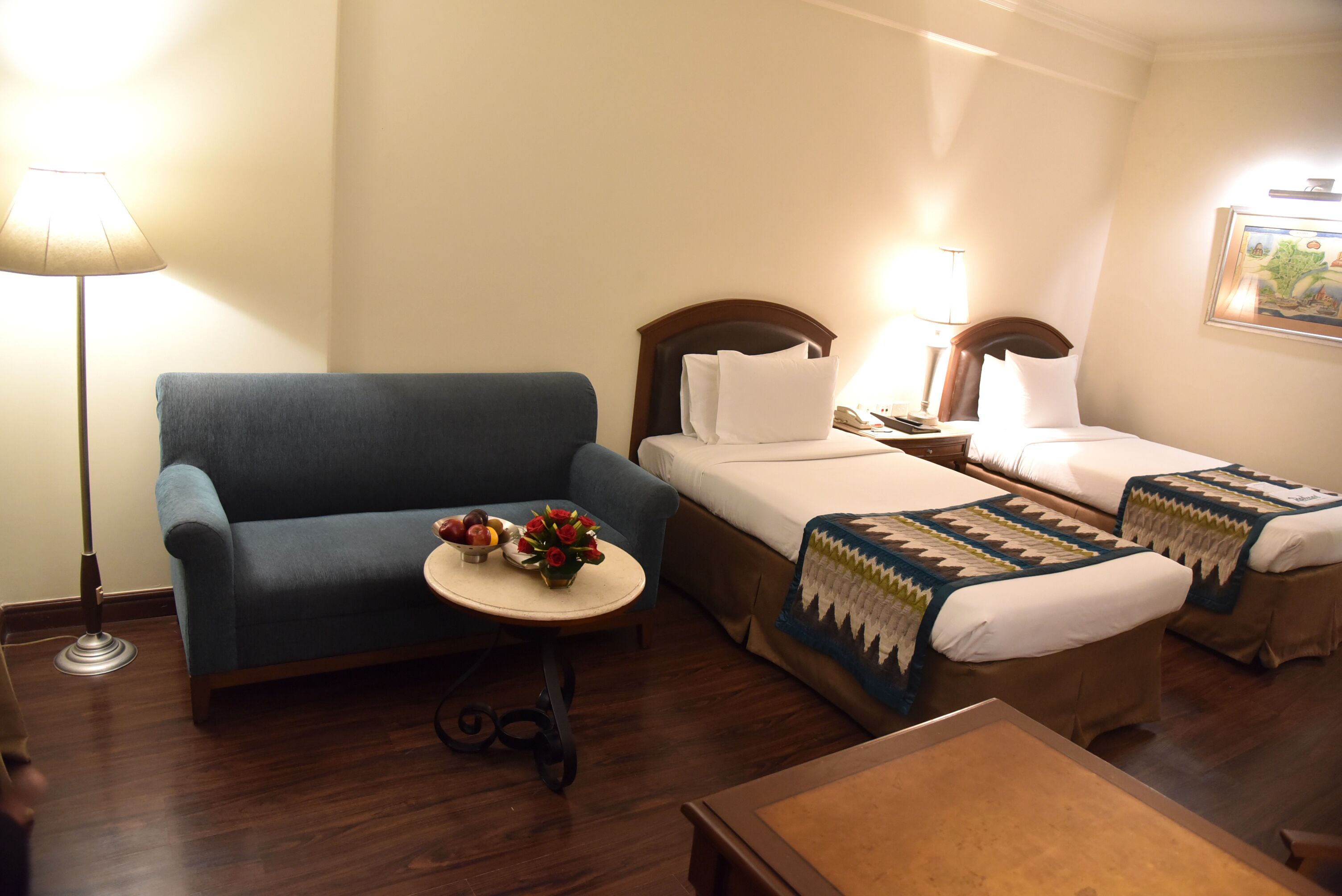 Habitación de hotel con dos camas, un sofá, una mesita con un jarrón de flores y una lámpara.