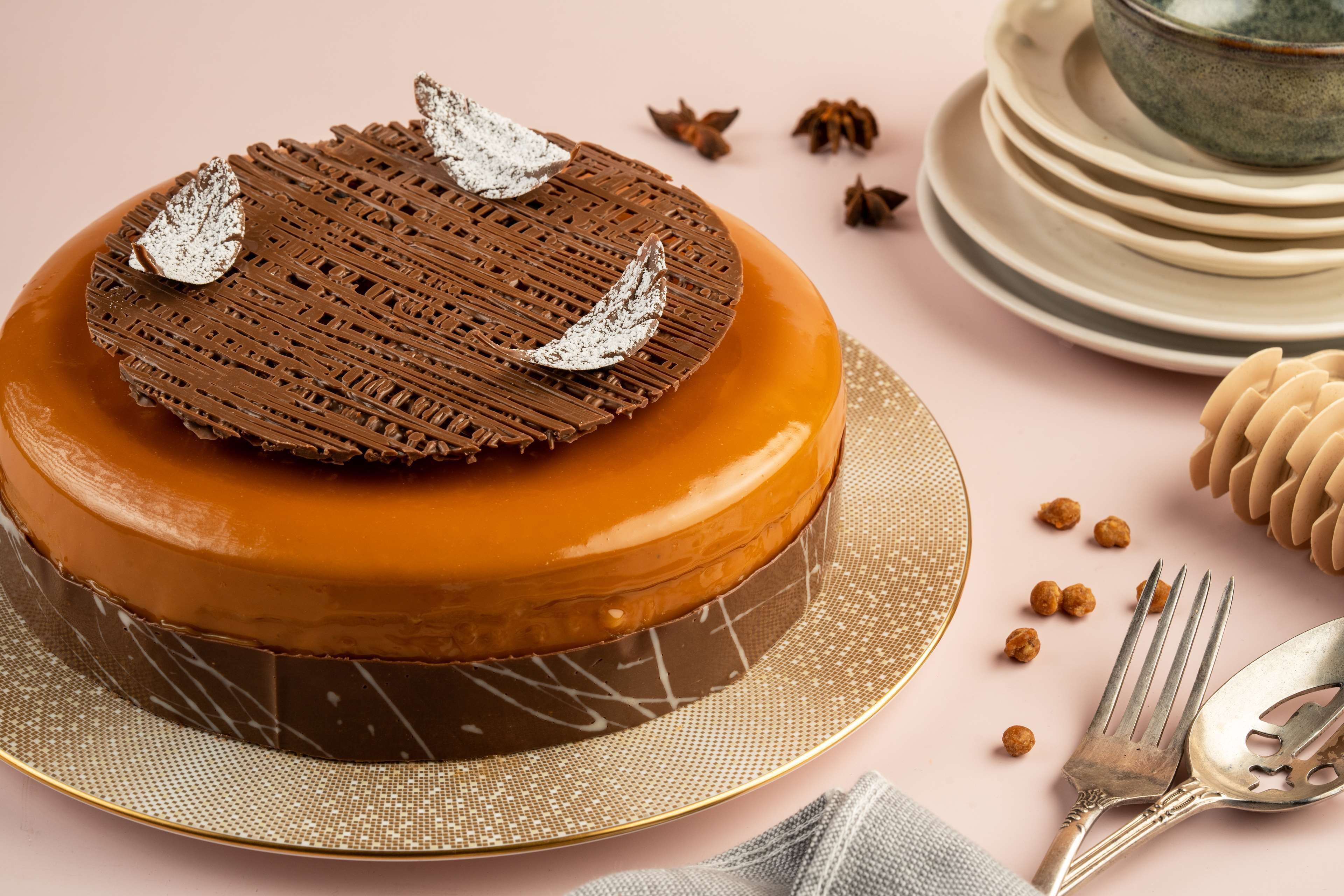 Un gâteau rond, surmonté d’un décor en treillis de chocolat et de feuilles argentées décoratives, posé sur un présentoir à gâteaux bordé d’or.