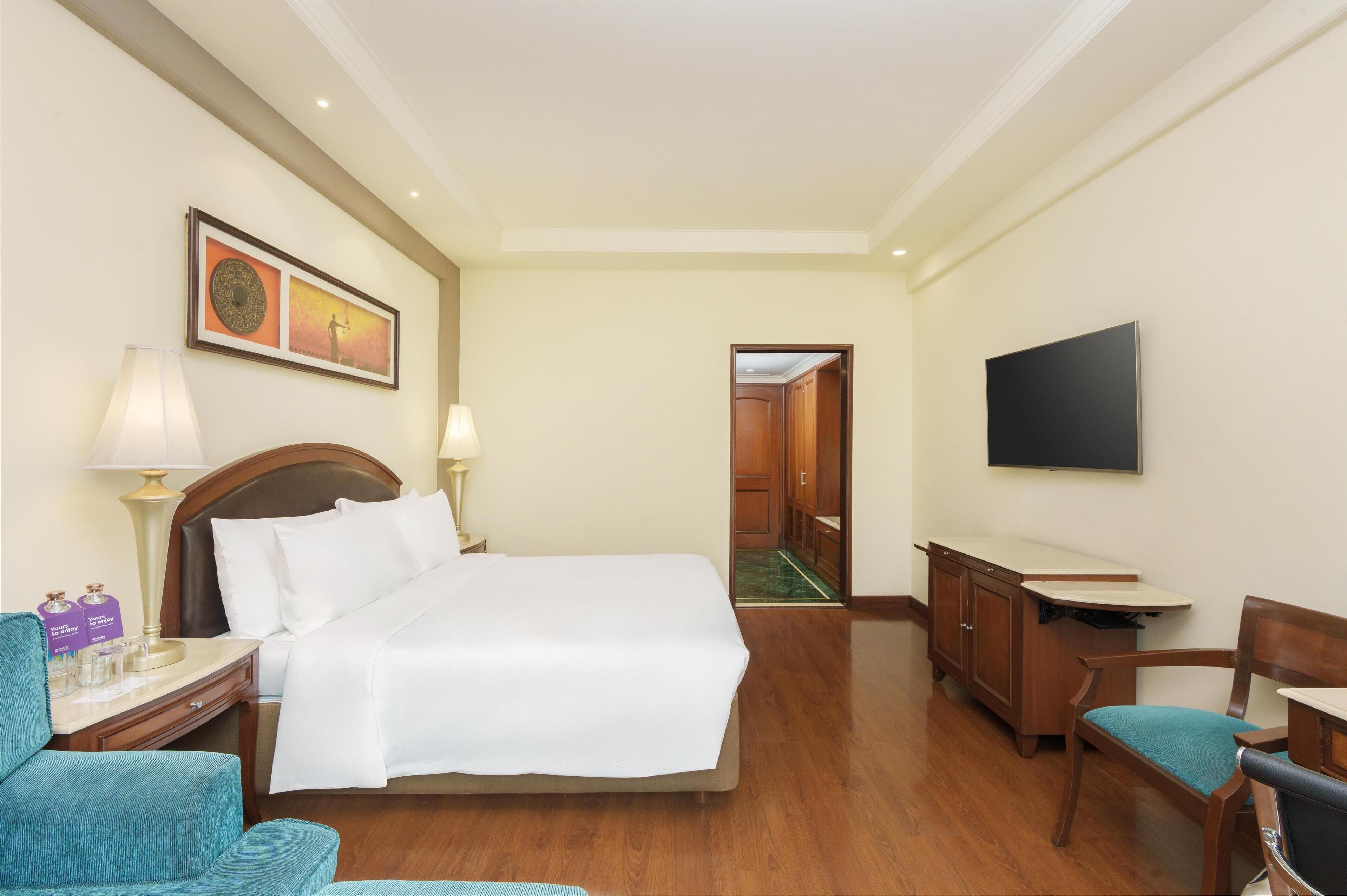 A hotel room with a bed, a desk, a chair, a television, and a painting on the wall.