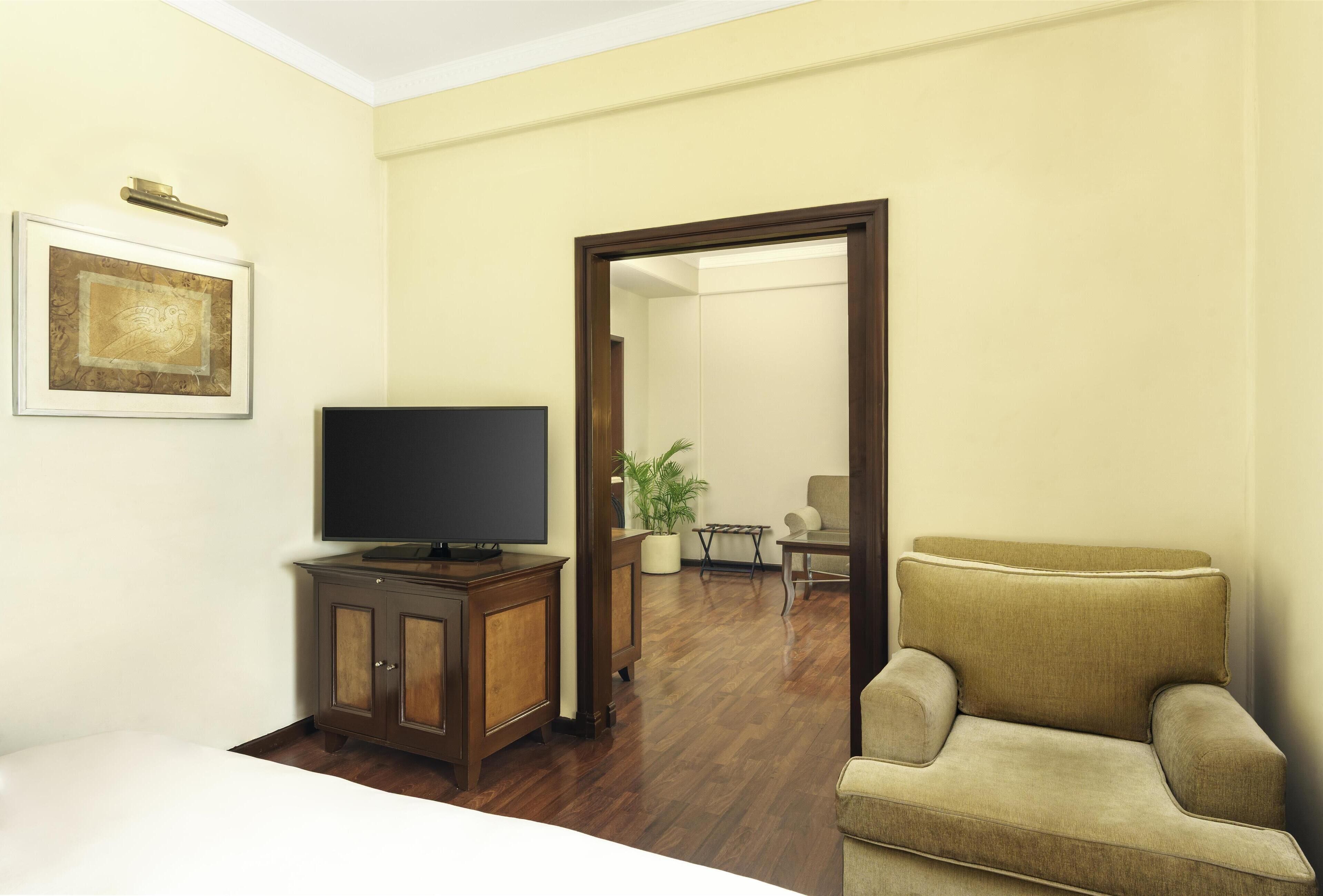 A hotel room with a bed, a TV on a wooden cabinet, a beige armchair, a framed picture on the wall, and a potted plant in the corner.
