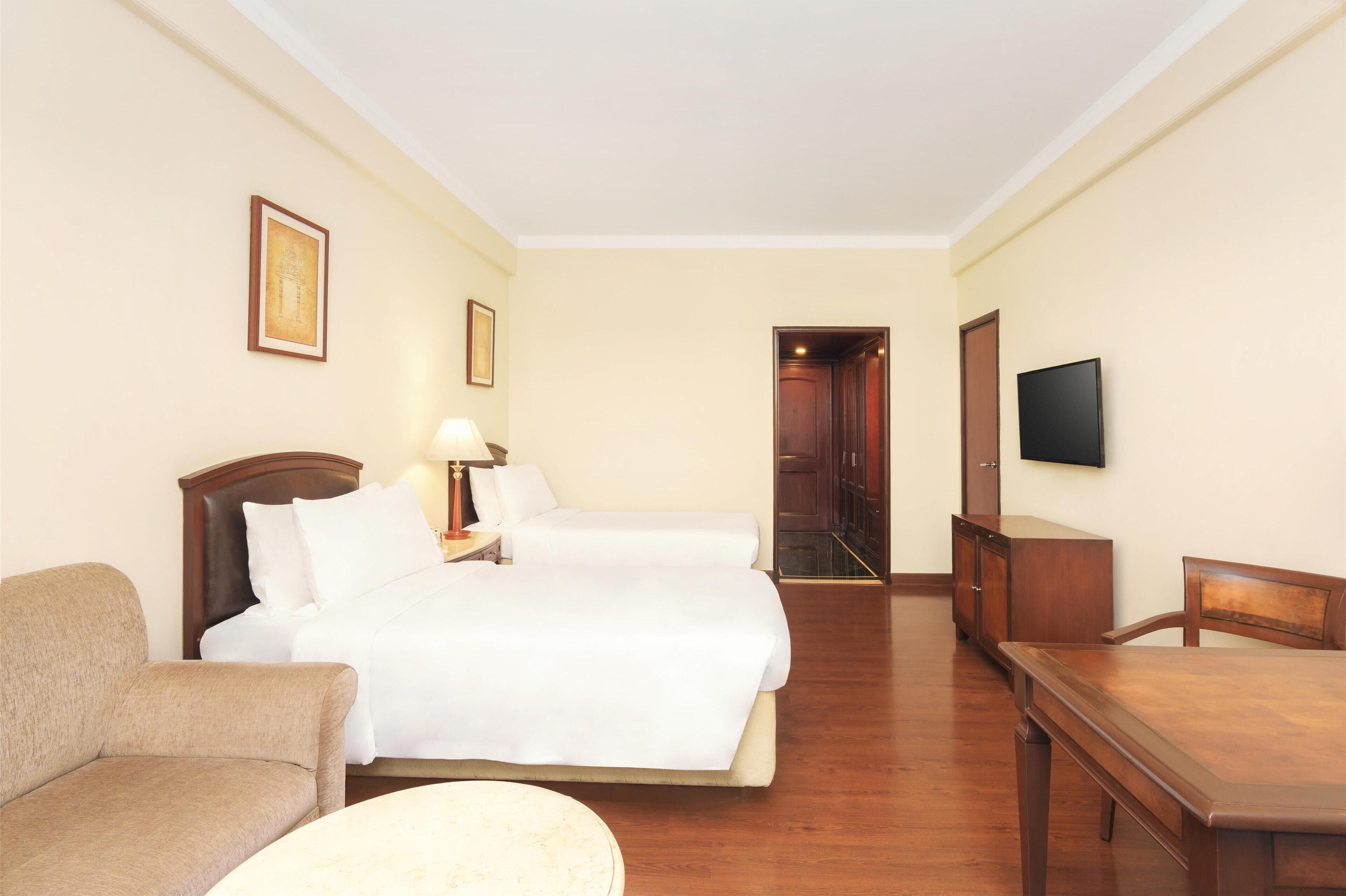 A hotel room with a bed, a chair, a table, a TV, and a door leading to a hallway.