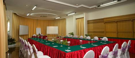 Une salle de conférence avec une longue table, des chaises blanches ornées de rubans violets et une nappe rouge.