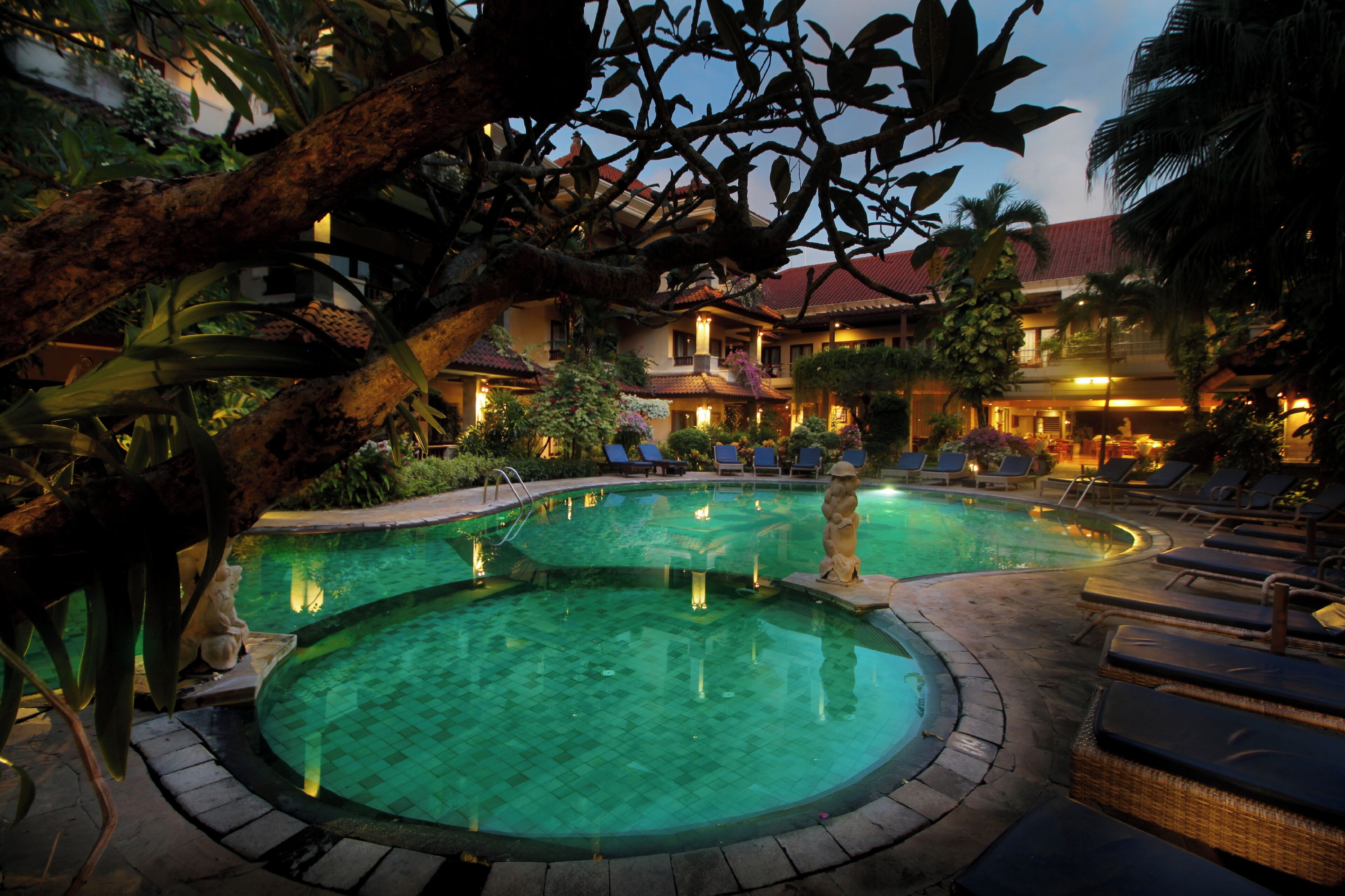 Un área de piscina con sillones de descanso y una estatua, rodeada de plantas tropicales y un edificio con tejado rojo.