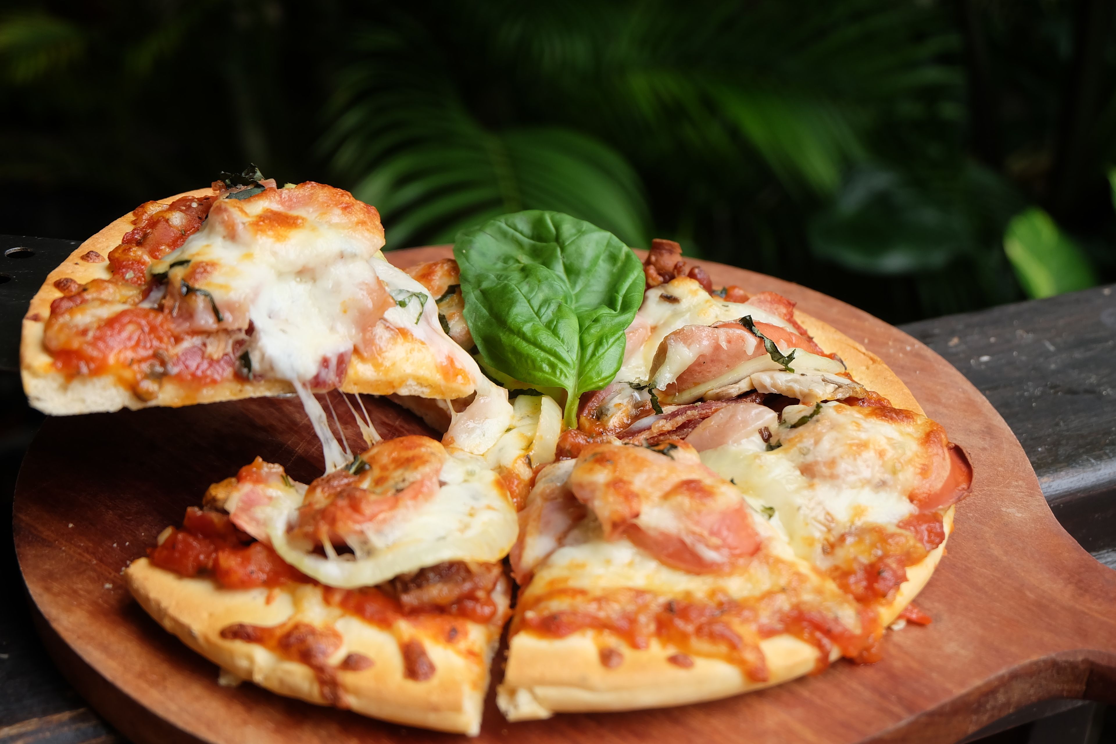Une pizza, posée sur une planche en bois, dont une part a été coupée, et qui est garnie de fromage, de tomates et de basilic.