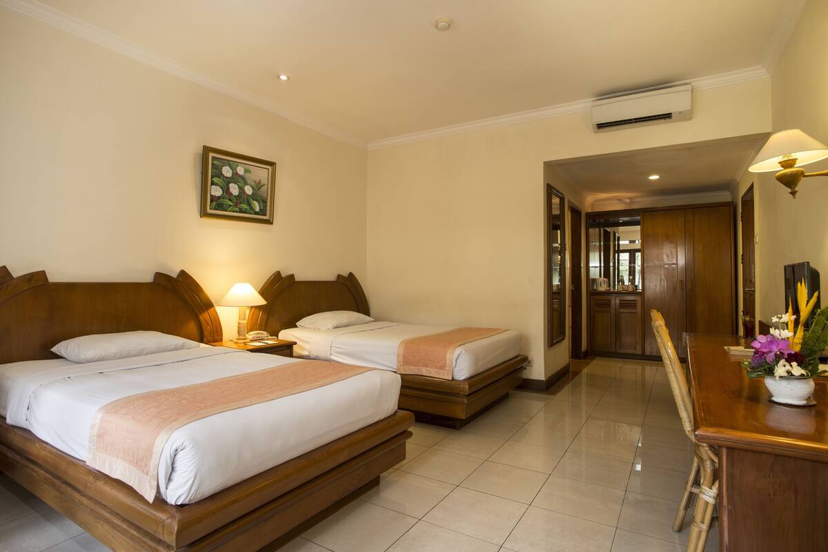 A hotel room with two beds, a wooden headboard, a bedside table with a lamp, a painting on the wall, and a bathroom visible through an open door.