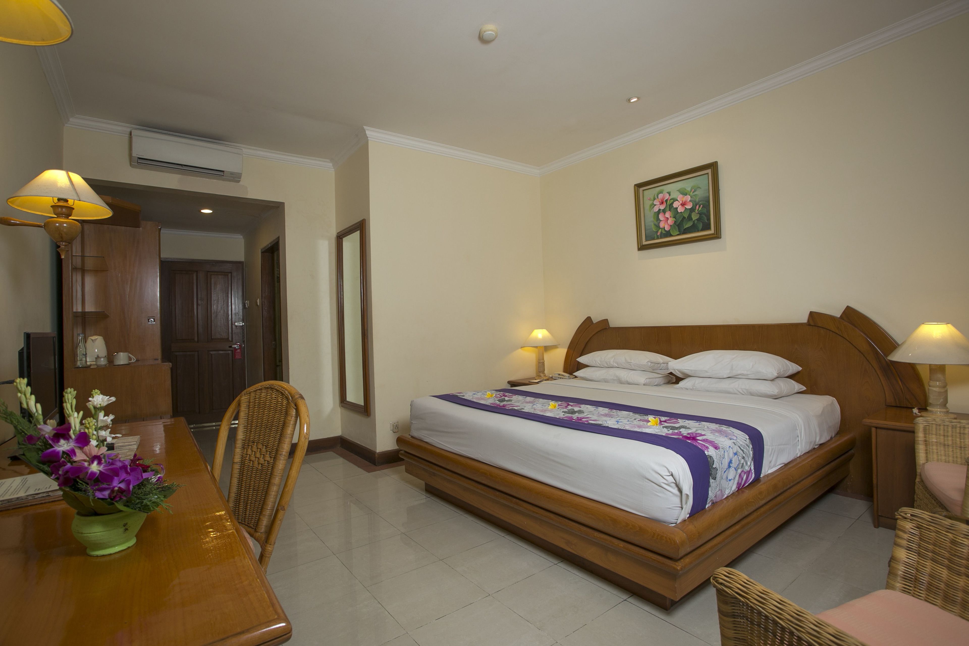 A hotel room with a bed, bedside tables, a desk with a chair, a dining table with chairs, and a flower arrangement.