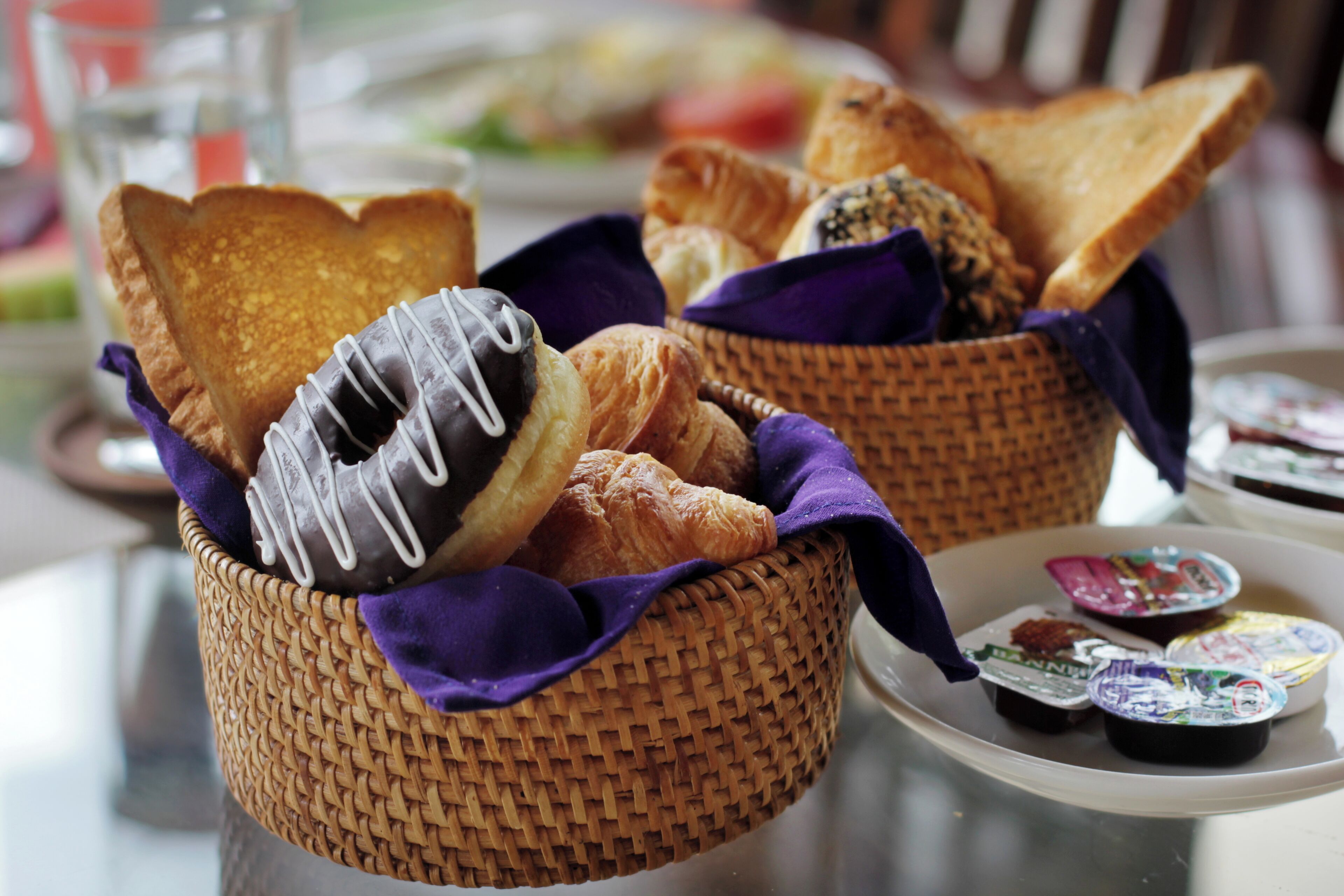 Un panier rempli d’une variété de viennoiseries et de pains, une assiette avec des pots de yaourt et un verre d’eau sur une table.