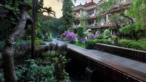 A lush garden with a waterfall, a wooden deck, and a multi-story building with balconies. - Parigata Resort & Spa (Denpasar)