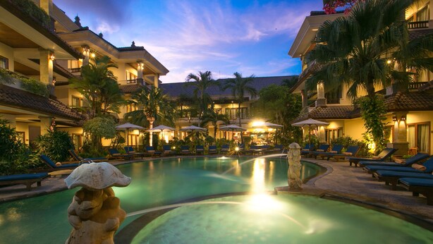 A hotel pool area with lounge chairs and umbrellas, surrounded by buildings with balconies and palm trees. - Parigata Resort & Spa (Denpasar)