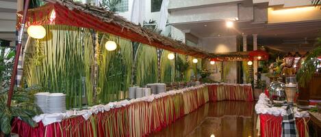 A long banquet table with a red and gold draped tablecloth, surrounded by tall green plants and hanging lights.