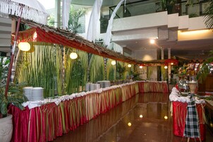 A long banquet table with a red and gold draped tablecloth, surrounded by tall green plants and hanging lights.