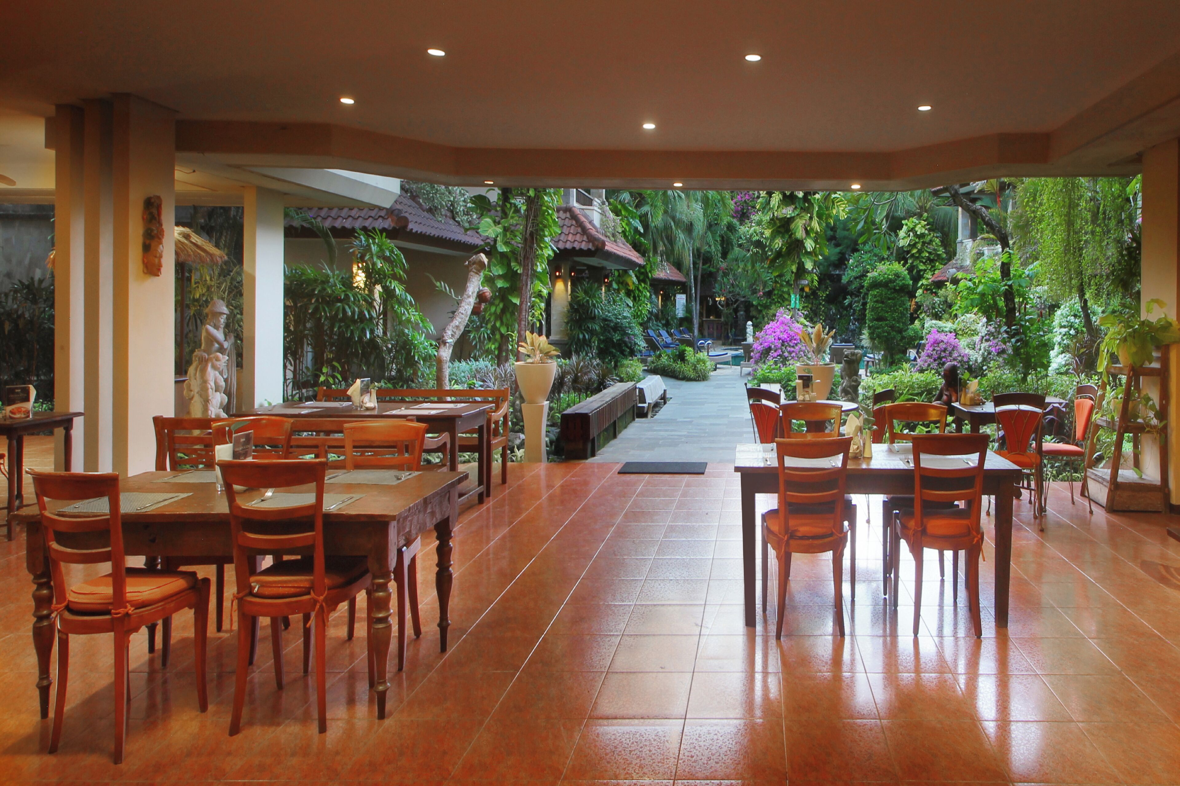 Comedor con mesas y sillas de madera, vista a un jardín exuberante con plantas diversas y un sendero que conduce al exterior.