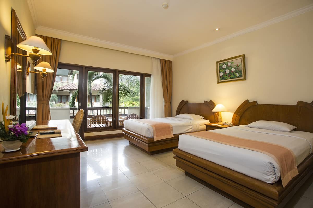A hotel room with two beds, a desk, and a balcony with a view of palm trees.
