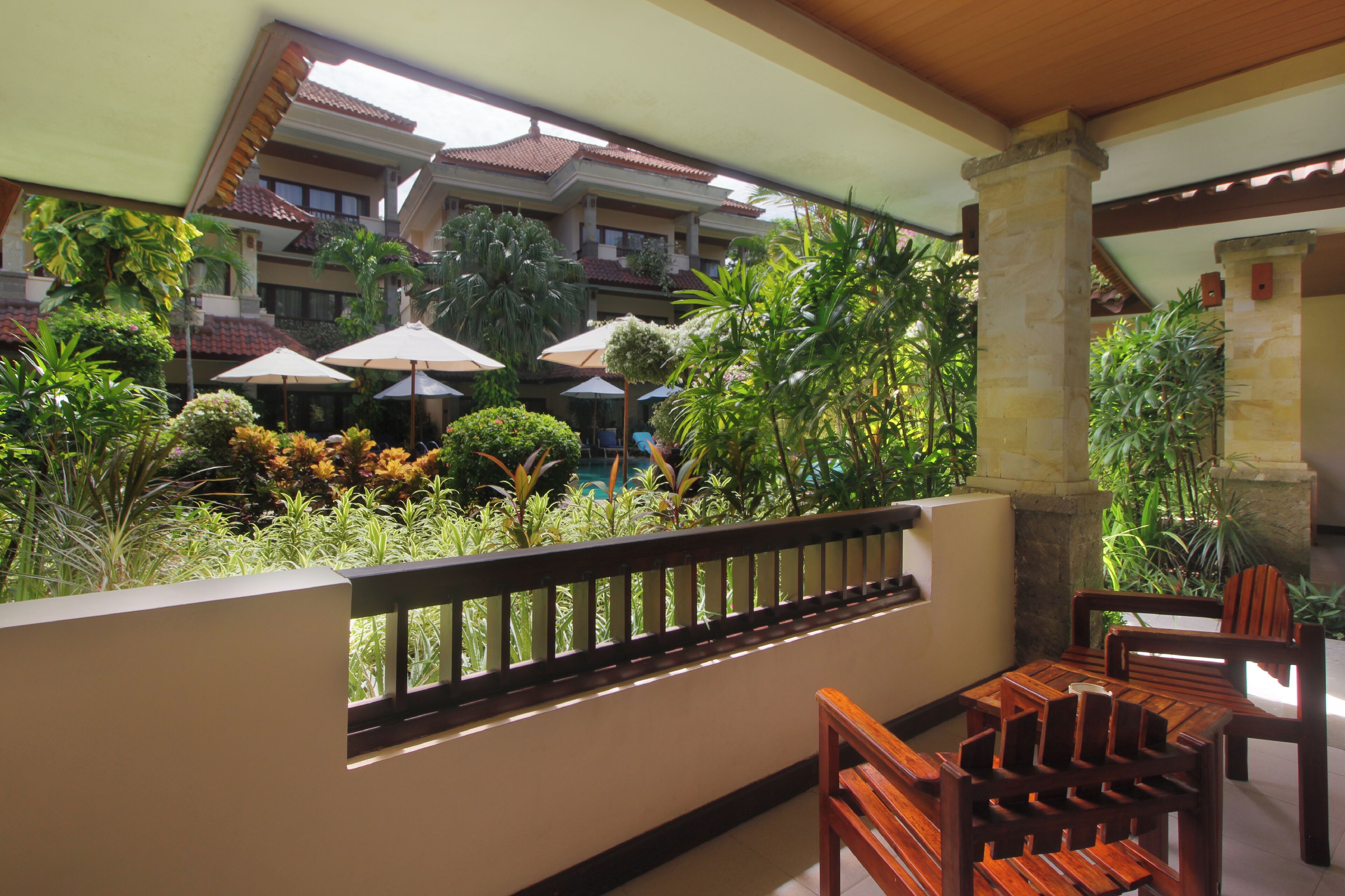 A balcony with wooden furniture and umbrellas, overlooking a lush garden and a building with a tiled roof.