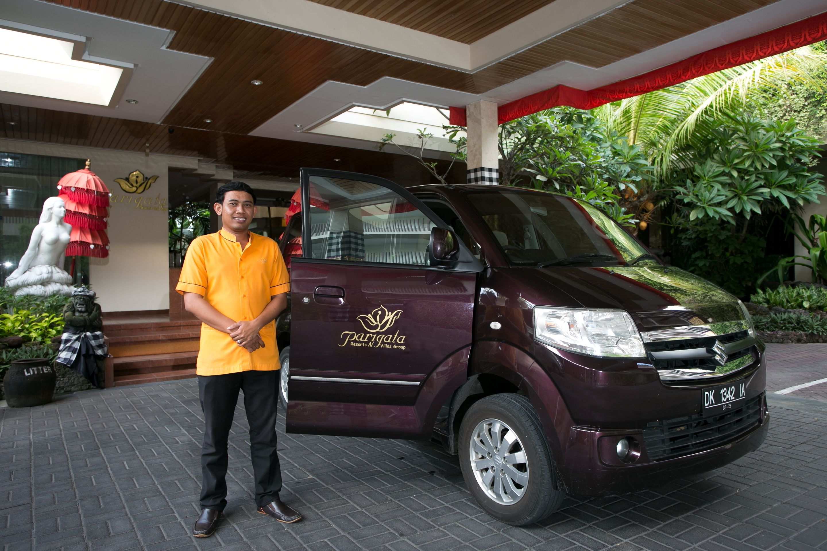 A man standing next to a maroon van with a logo on the side, parked in front of a building with a red awning and greenery.
