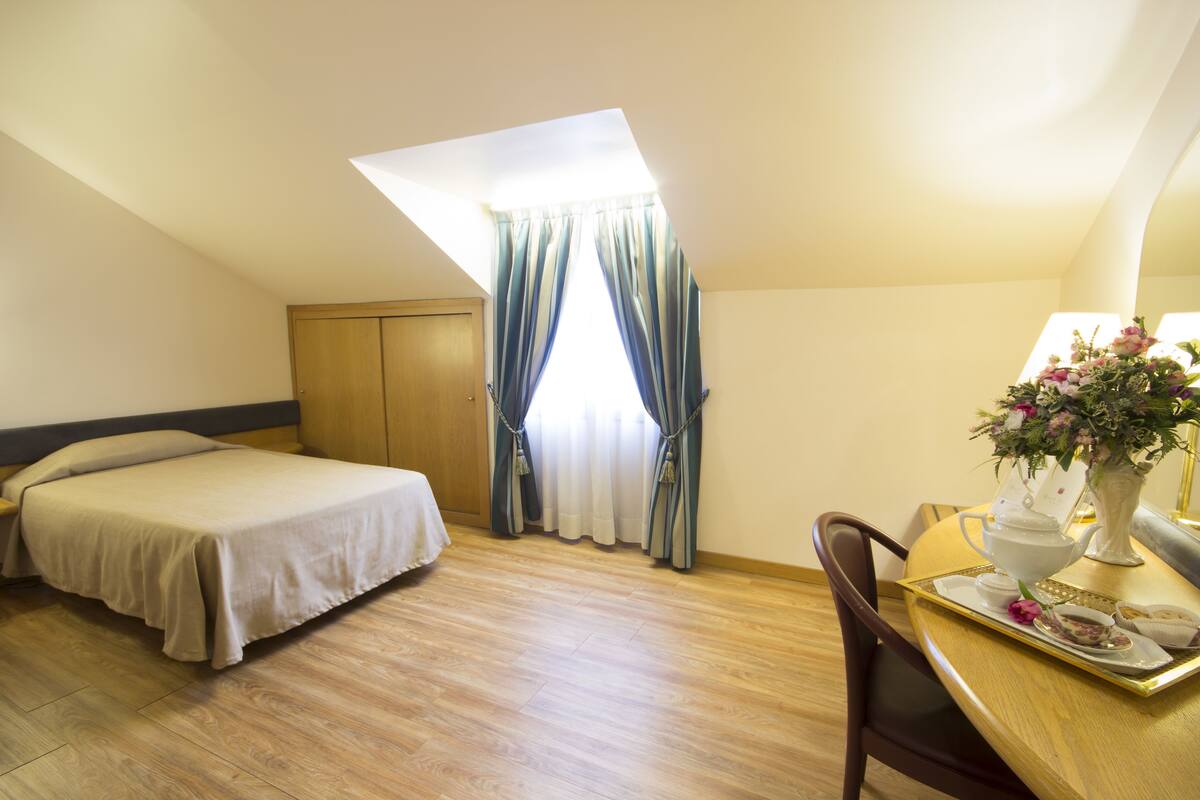 A room with a bed, a dining table with a tray, a vase with flowers, and a window with curtains.