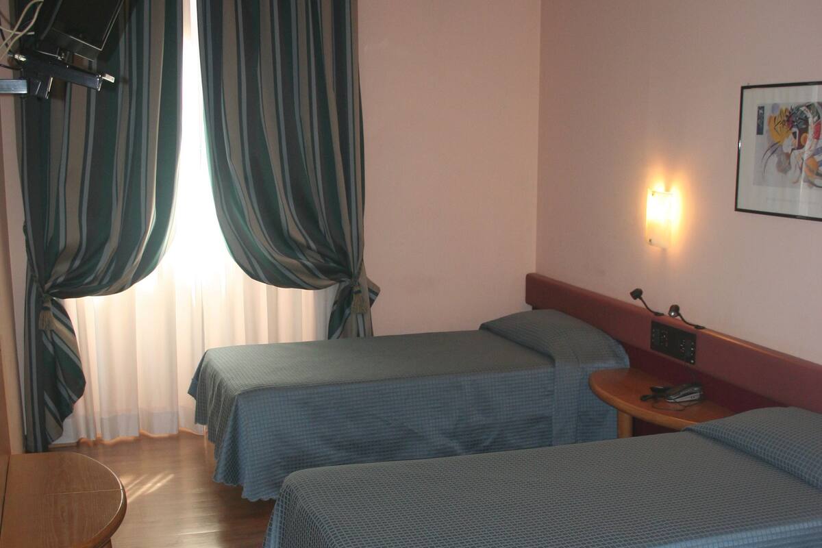 A hotel room with two single beds, a wooden table, a chair, and a window with curtains.