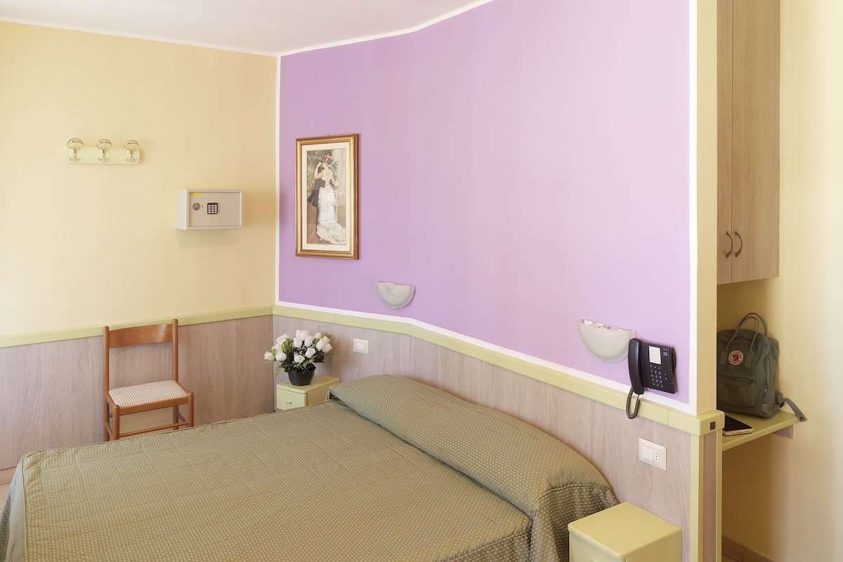 A bedroom with a bed, a chair, a bedside table with a lamp, a wall-mounted phone, and a framed picture.