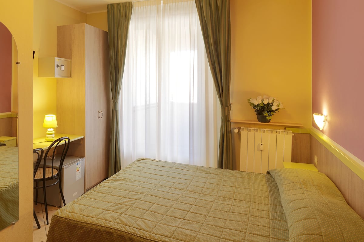 A hotel room with a bed, a desk, a chair, a radiator, and a potted plant.