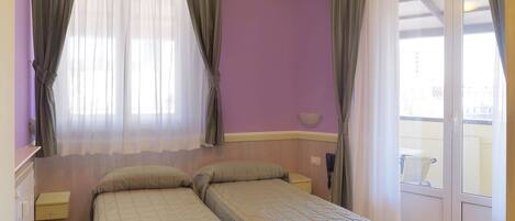 A room with two beds, a window with sheer curtains, a small table, and a view of the outside.