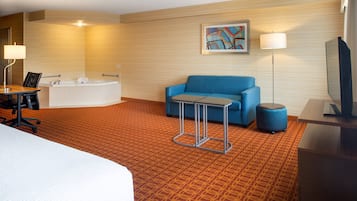A hotel room with a large bed, a sofa, a small table, a lamp, a TV, a bathtub, and a painting on the wall.