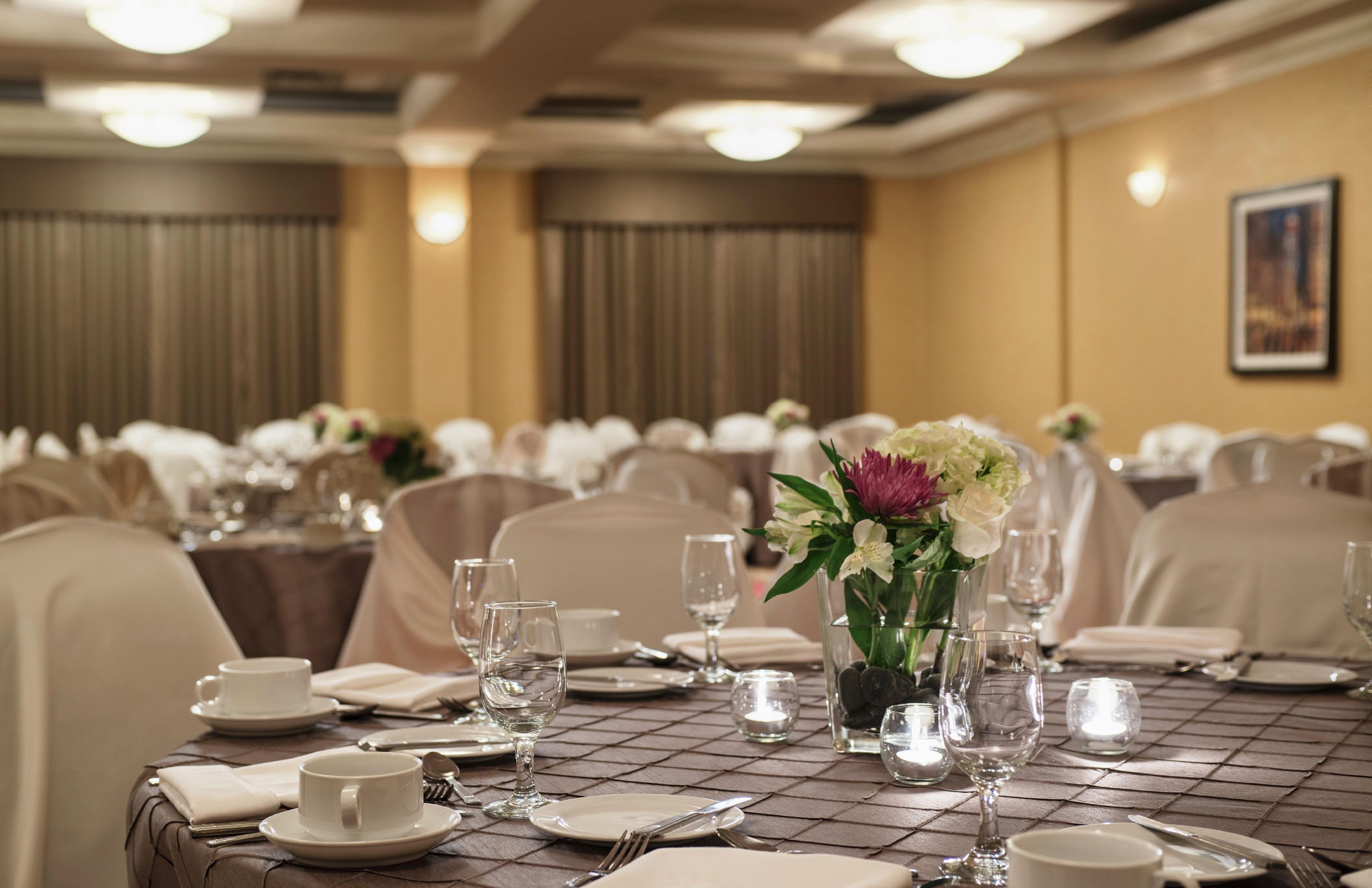 Une salle de banquet où les tables sont dressées pour le repas, avec des nappes blanches, de la verrerie et des compositions florales en guise de centre de table.