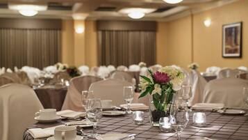 Une salle de banquet où les tables sont dressées pour le repas, avec des nappes blanches, de la verrerie et des compositions florales en guise de centre de table.
