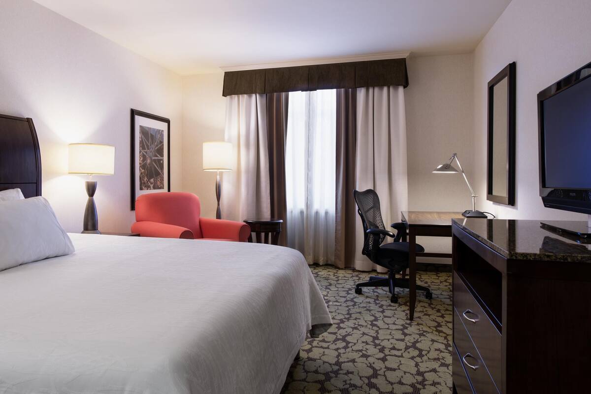 A hotel room with a bed, a desk with a chair, a TV, and a window with curtains.