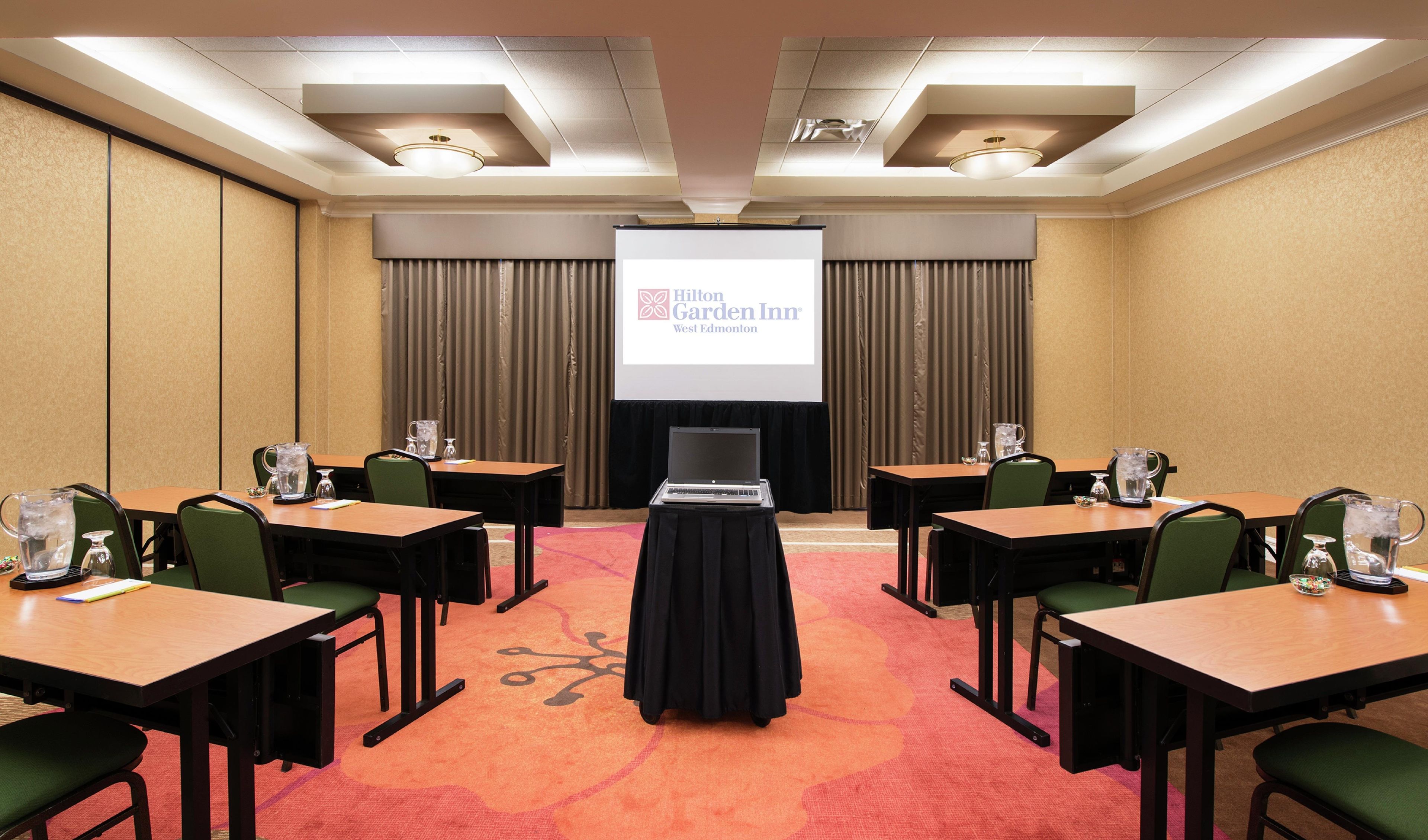 Un salón de conferencias con una pantalla de proyector mostrando el logotipo de Hilton Garden Inn West Edmonton, varias mesas con sillas y un ordenador portátil sobre un soporte.