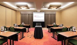 Un salón de conferencias con una pantalla de proyector mostrando el logotipo de Hilton Garden Inn West Edmonton, varias mesas con sillas y un ordenador portátil sobre un soporte.