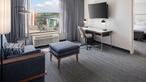 A hotel room with a large window, a desk, a chair, a TV, and a bed.