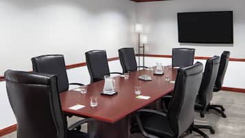 A conference room with a long table, eight black leather chairs, a flat-screen TV mounted on the wall, and a floor lamp.