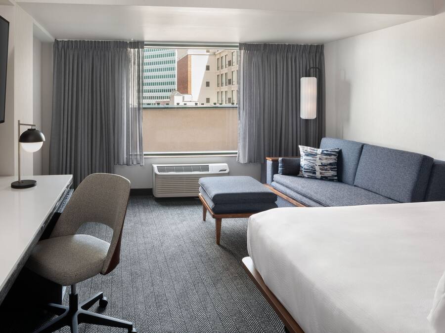 A hotel room with a bed, a desk, a chair, a sofa, and a large window with curtains.