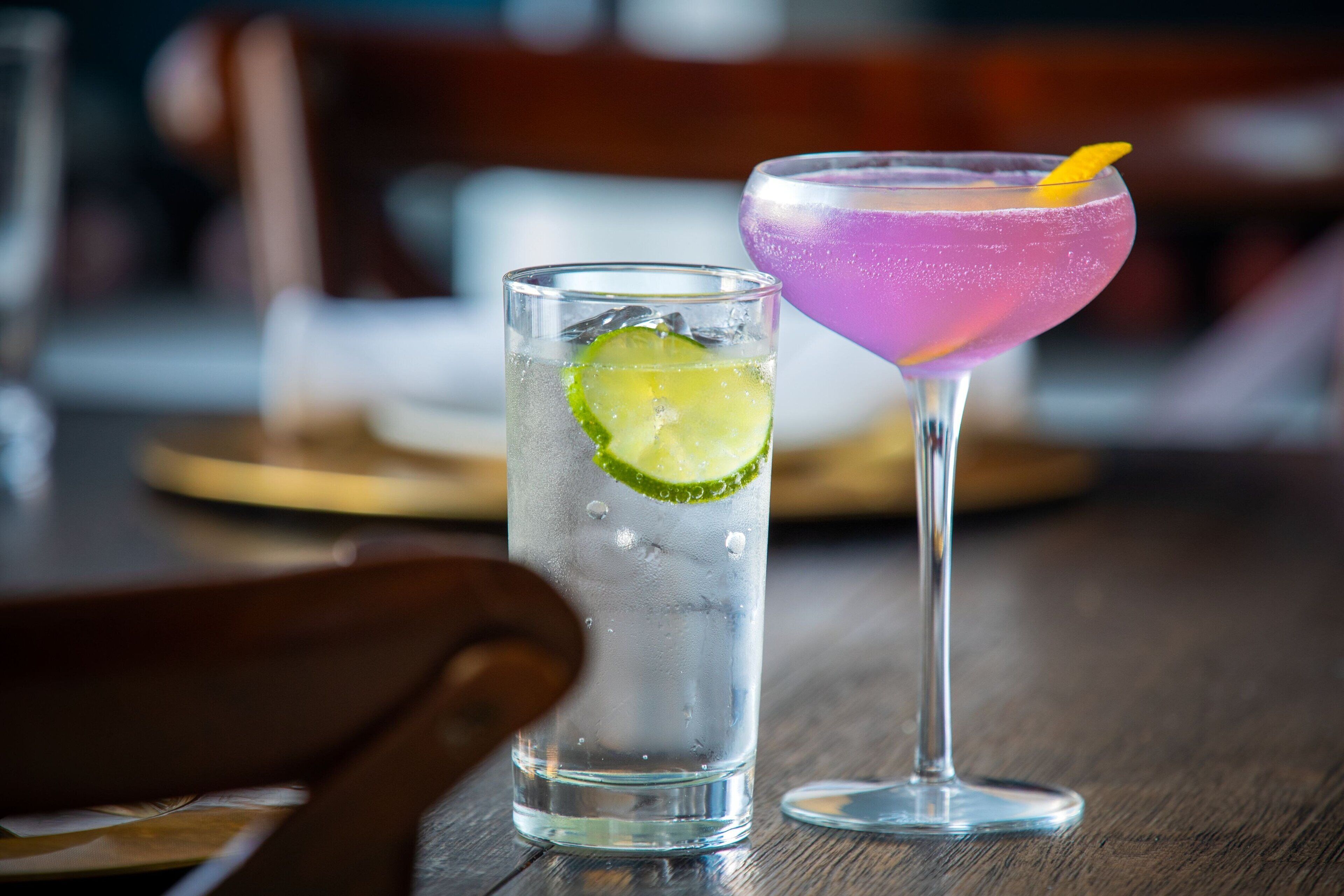 Two drinks on a wooden table, one in a tall glass and one in a martini glass, with a lime wedge in the tall glass.