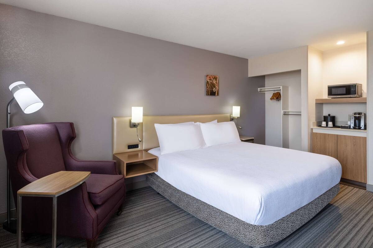 A hotel room with a bed, a chair, a desk, a nightstand, a lamp, a wall art piece, and a kitchenette with a microwave.